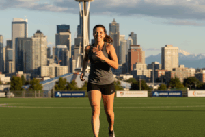 A smiling woman running on a sports field with the Seattle skyline in the background, symbolizing successful ACL rehab and return to sport in Seattle physical therapy.